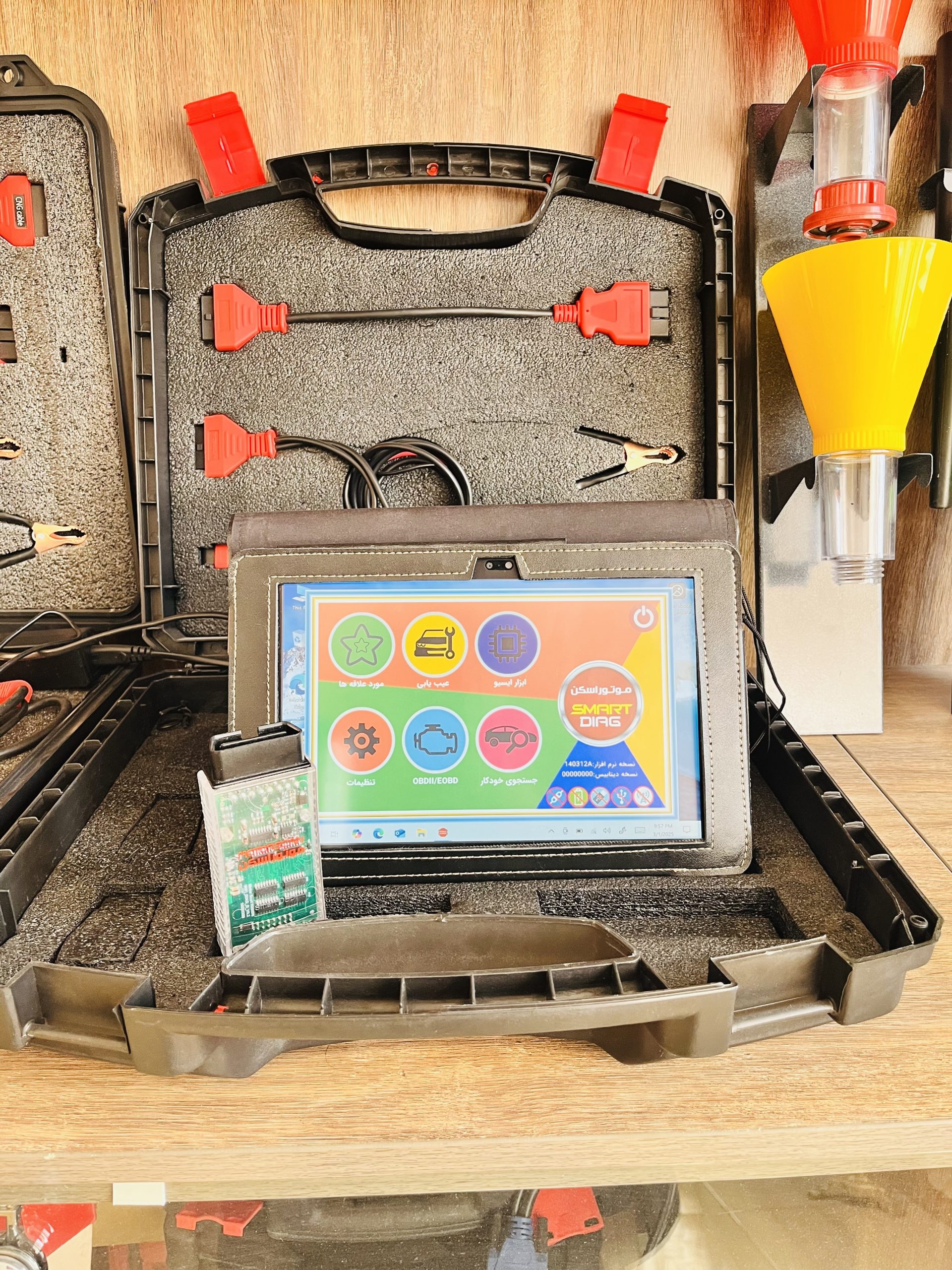 A compact black plastic case with a custom foam interior holds a collection of red and black cables, connectors, and a transparent electronic module. In the center lies a tablet with a vibrant, colorful interface displaying Arabic text and icons representing various functions like diagnostics and settings. To the side of the case, a yellow funnel-like device mounted on a stand is visible. The setup seems to be part of a professional-grade automotive diagnostic toolkit, designed for both functionality and ease of use